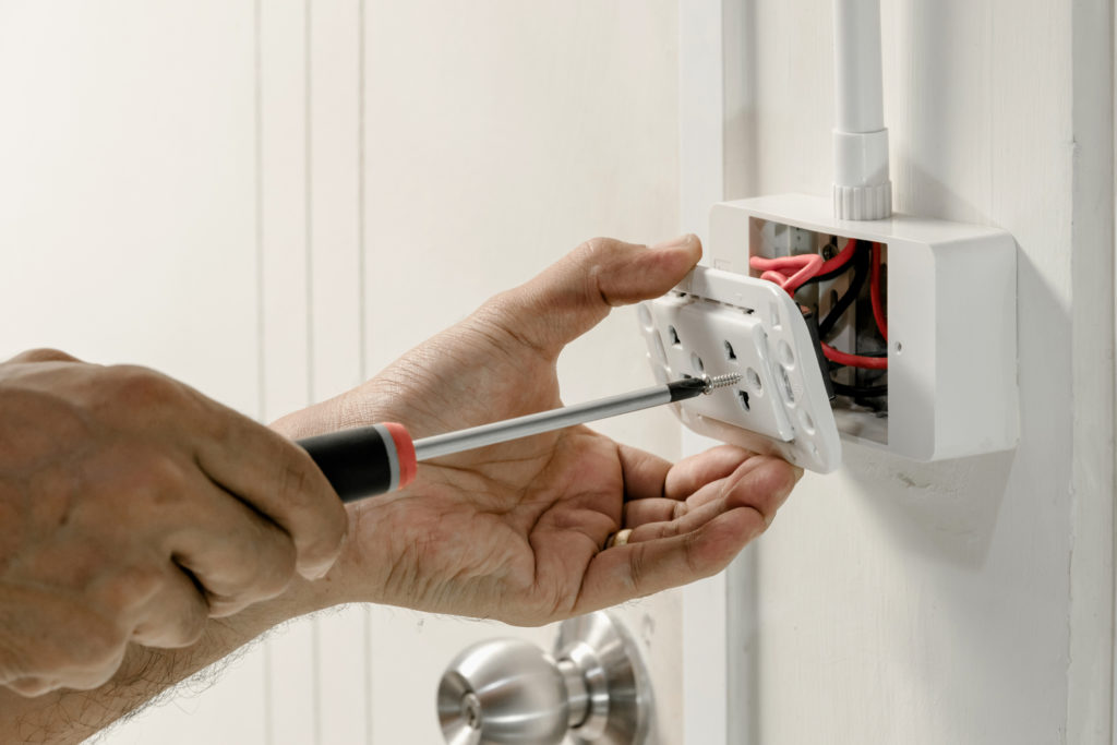Pair of hands screwing on an electrical outlet to a white wall. Wiring visible behind plate.